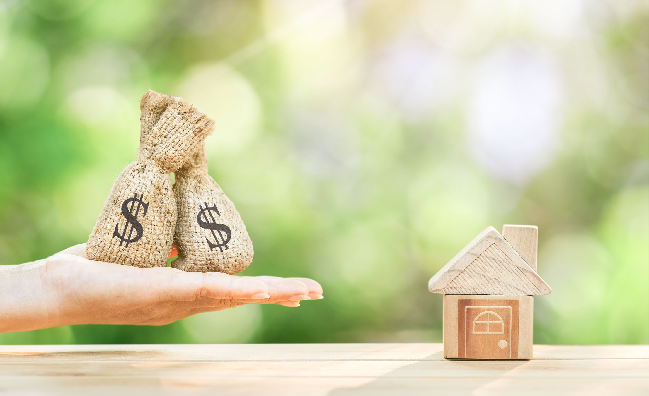 Hand holding money bags next to a small wooden toy house
